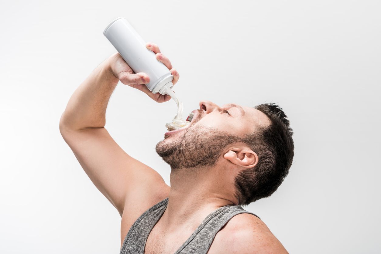chubby man in grey tank top spraying whipped cream in mouth on white