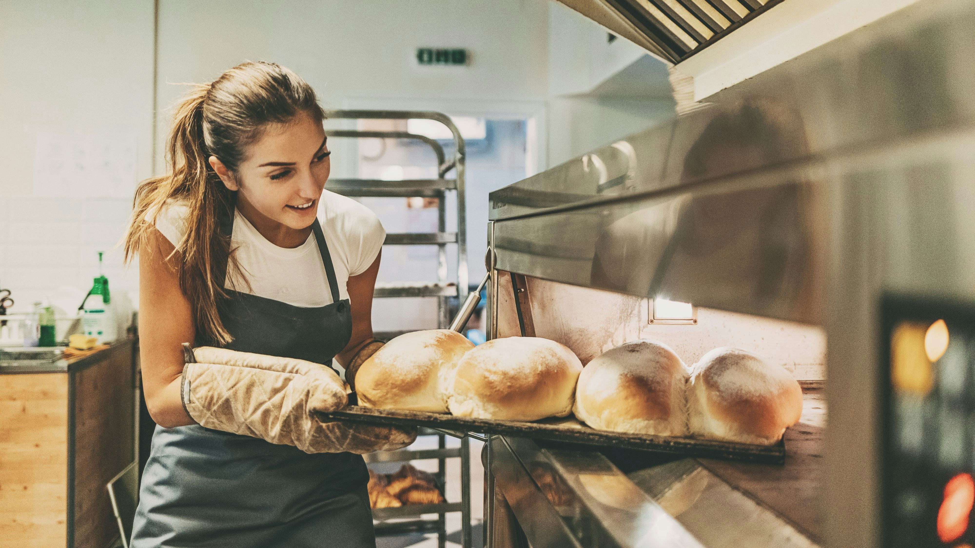 Young woman baker taking out the hot bread from the oven.