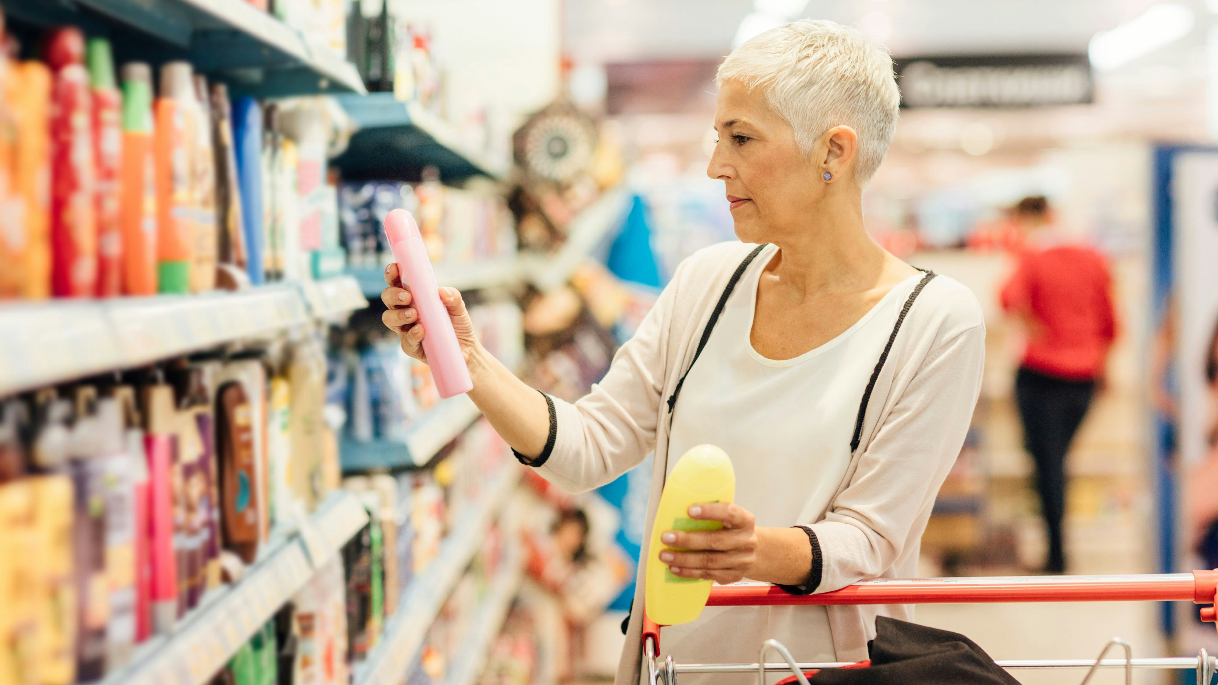 Mature smiling woman shopping in local supermarket. Standing by produce stand and choosing cosmetics, choosing shampoos.