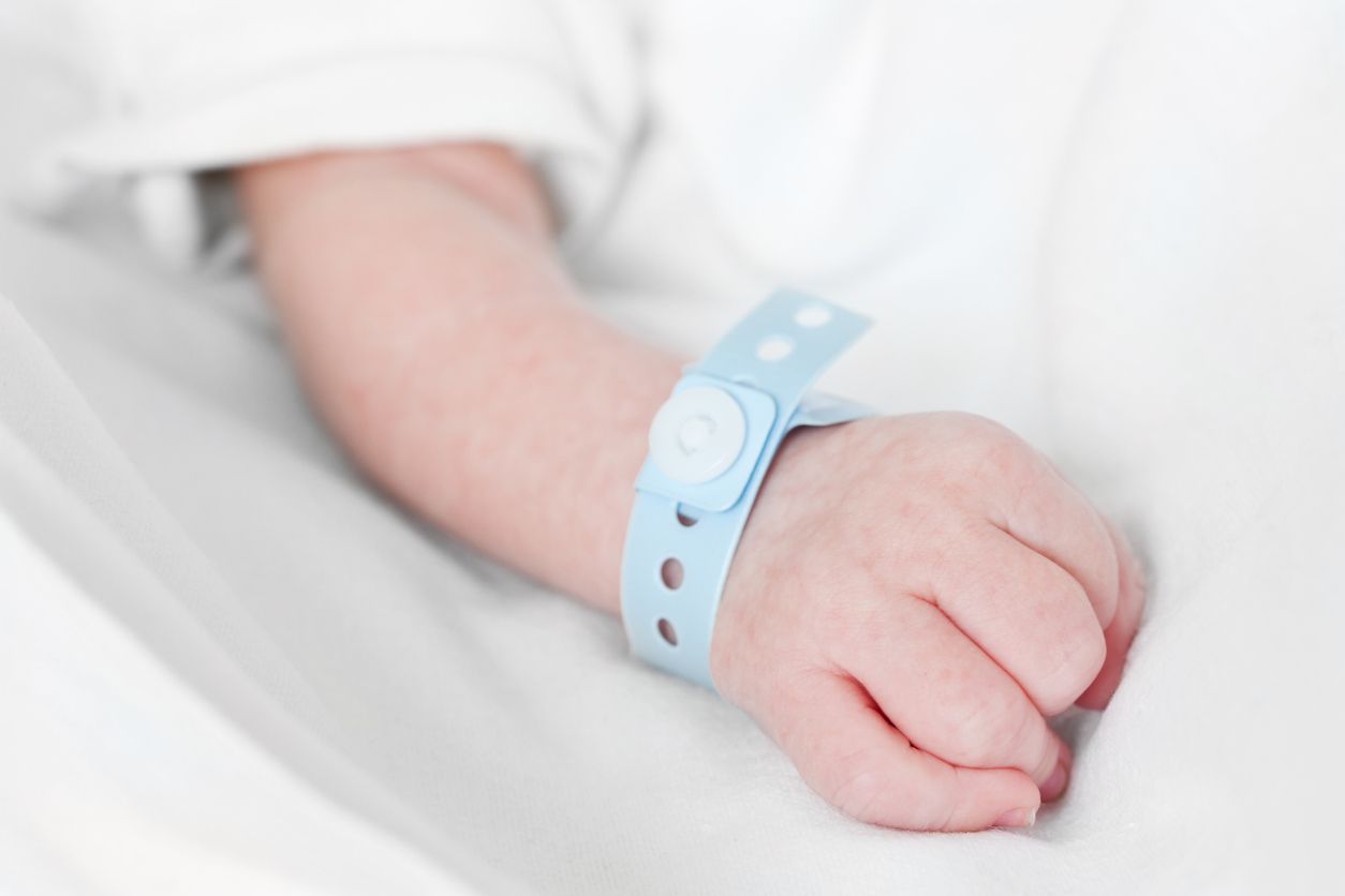 Close-up of a fist of a caucasian newborn baby with a hospital nametag attached to the wrist on white linen