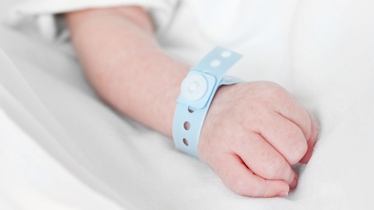 Close-up of a fist of a caucasian newborn baby with a hospital nametag attached to the wrist on white linen