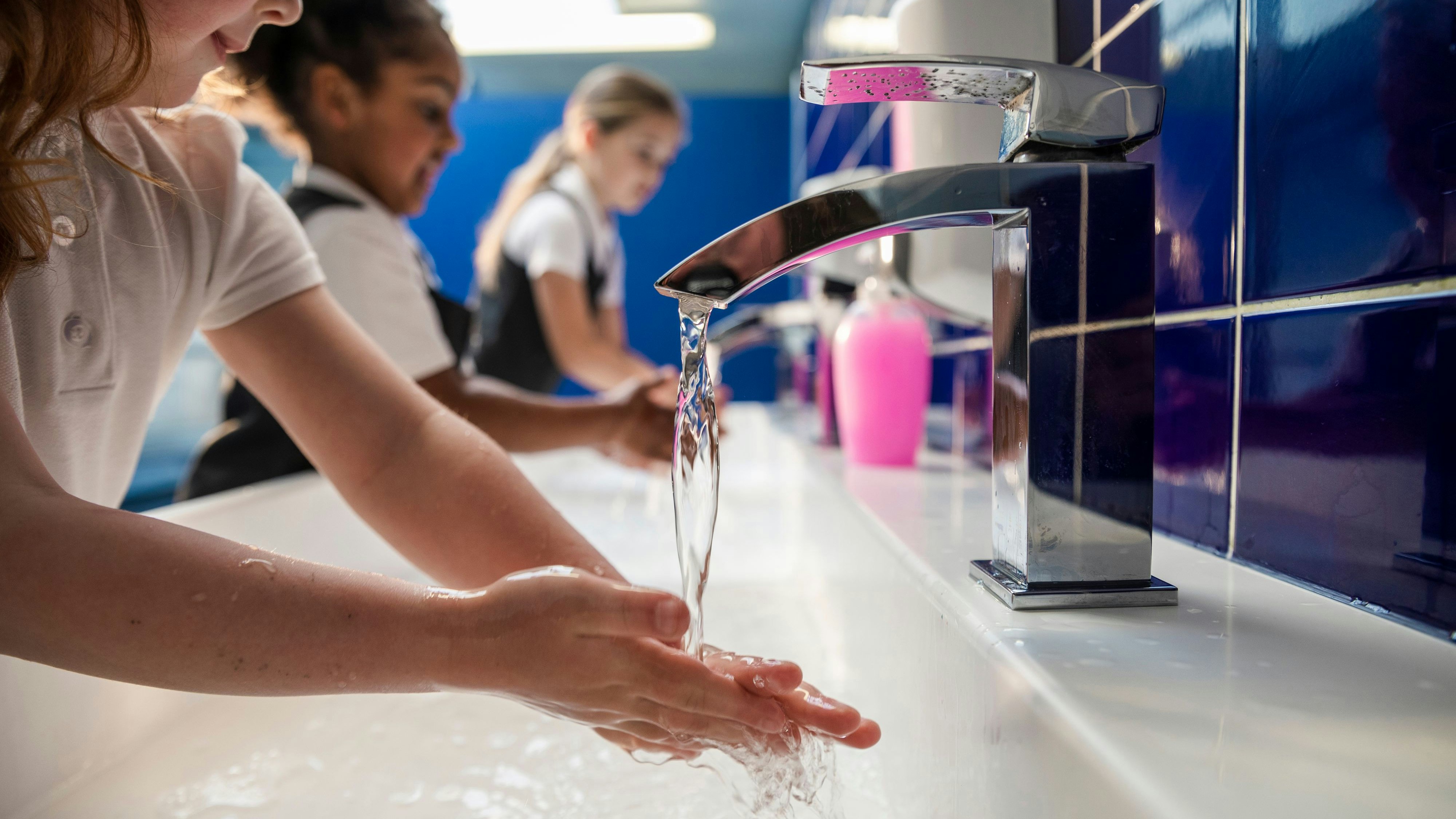 A side-view of diverse UK young schoolgirls, going through COVID-protocols and washing their hands with soap and water in their school toilets. They are all wearing their school uniforms.