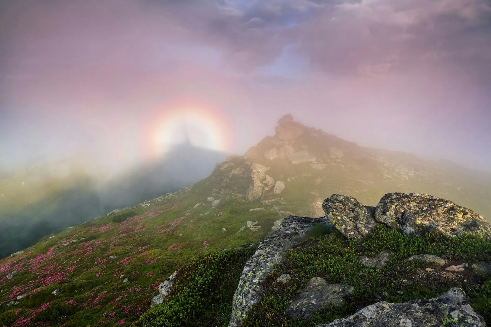 Wer auf einem Grat oder Gipfel mit dem Rücken zur Sonne steht, kann manchmal in der Ferne eine unheimliche Gestalt beobachten, die von einem leuchtenden Regenbogen-Ring umhüllt ist.