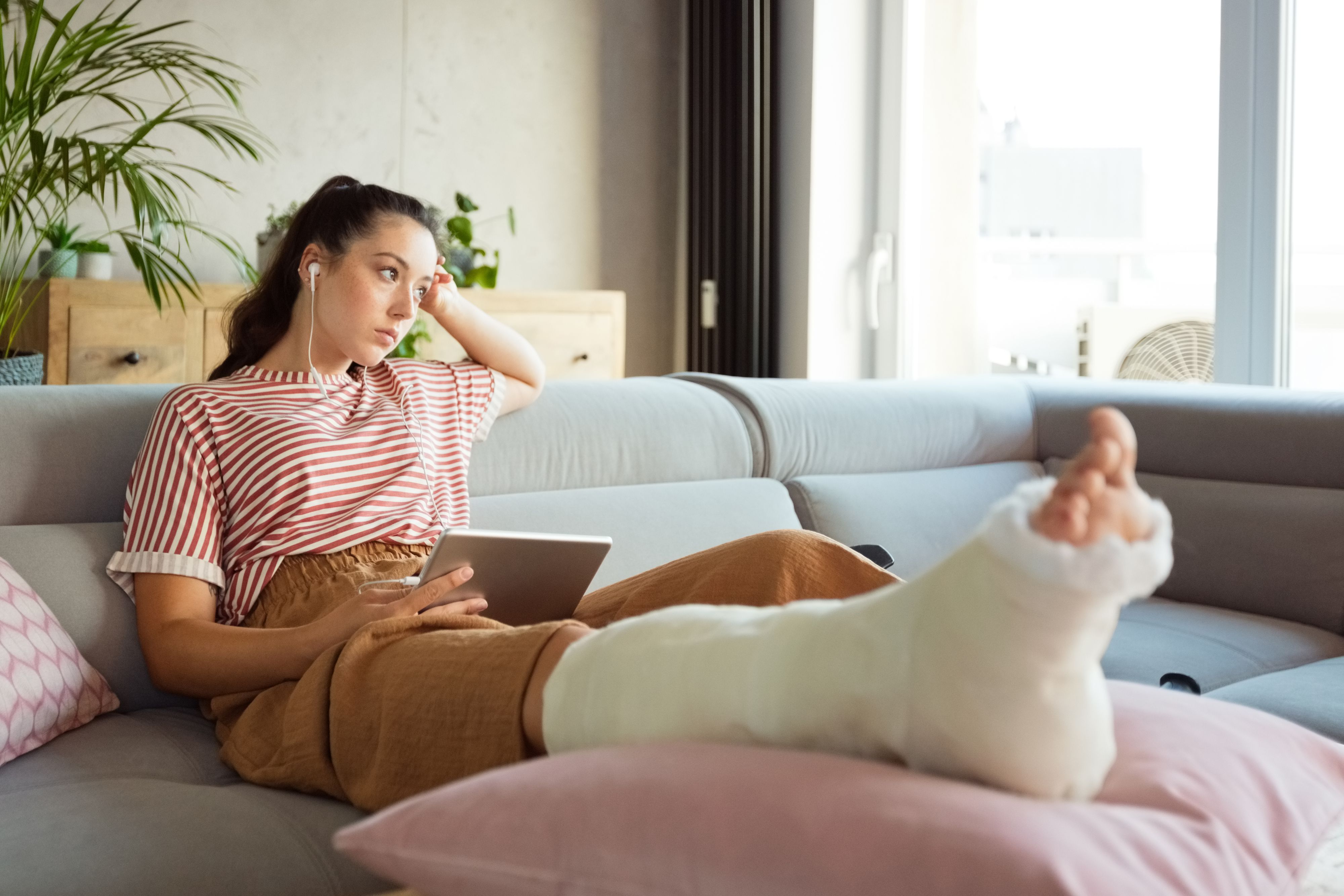 Worried young man with broken leg in plaster cast lying down on sofa at home, holding a digital tablet and looking through window.