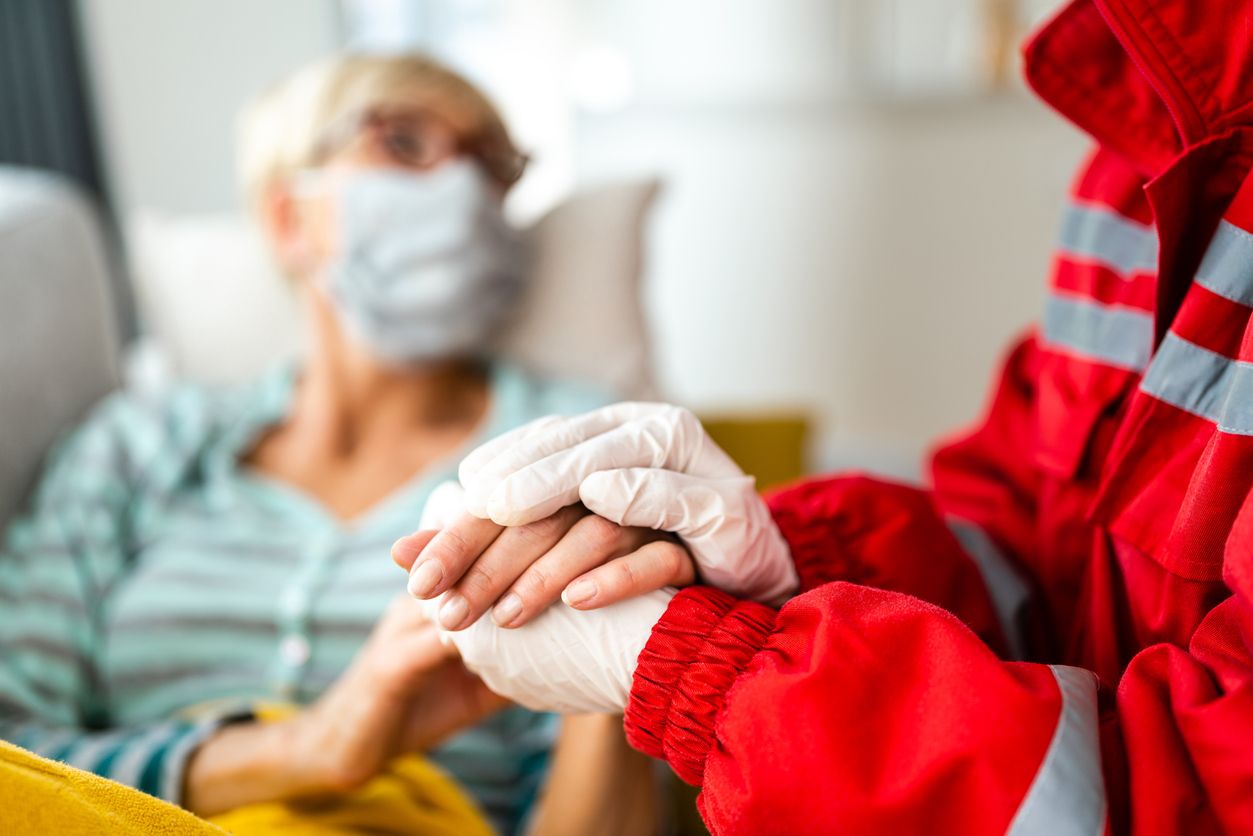 Close up of paramedic doctor holding hands with senior patient at home. Both of them are wearing protective face mask because of COVID-19.