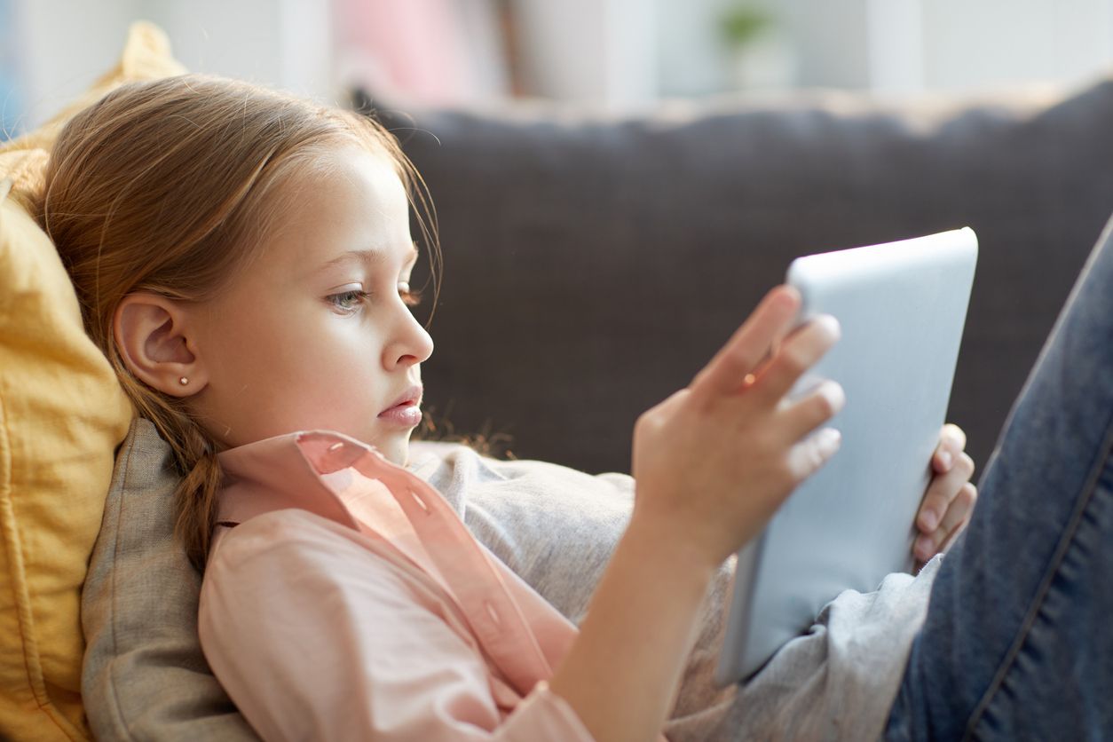 Side view portrait of little girl using digital tablet while lying on sofa, watching cartoons or browsing internet, copy space