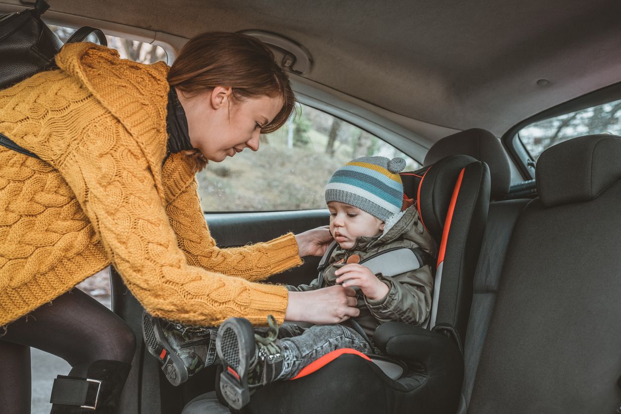 Photo of Young mother putting her little baby boy in the car seat, fastening seat belts during winter day. Protection in the car. Safe transportation of Toddler, children in the car. Brown hair female fastens the baby car seat. The baby is serious and looking away.