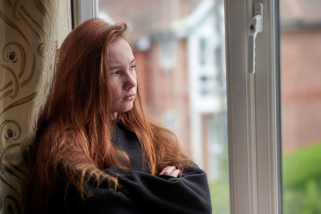 Side view of moody female teenager in black casual wear with red hair standing at window with arms crossed and looking away while staying at home under isolation