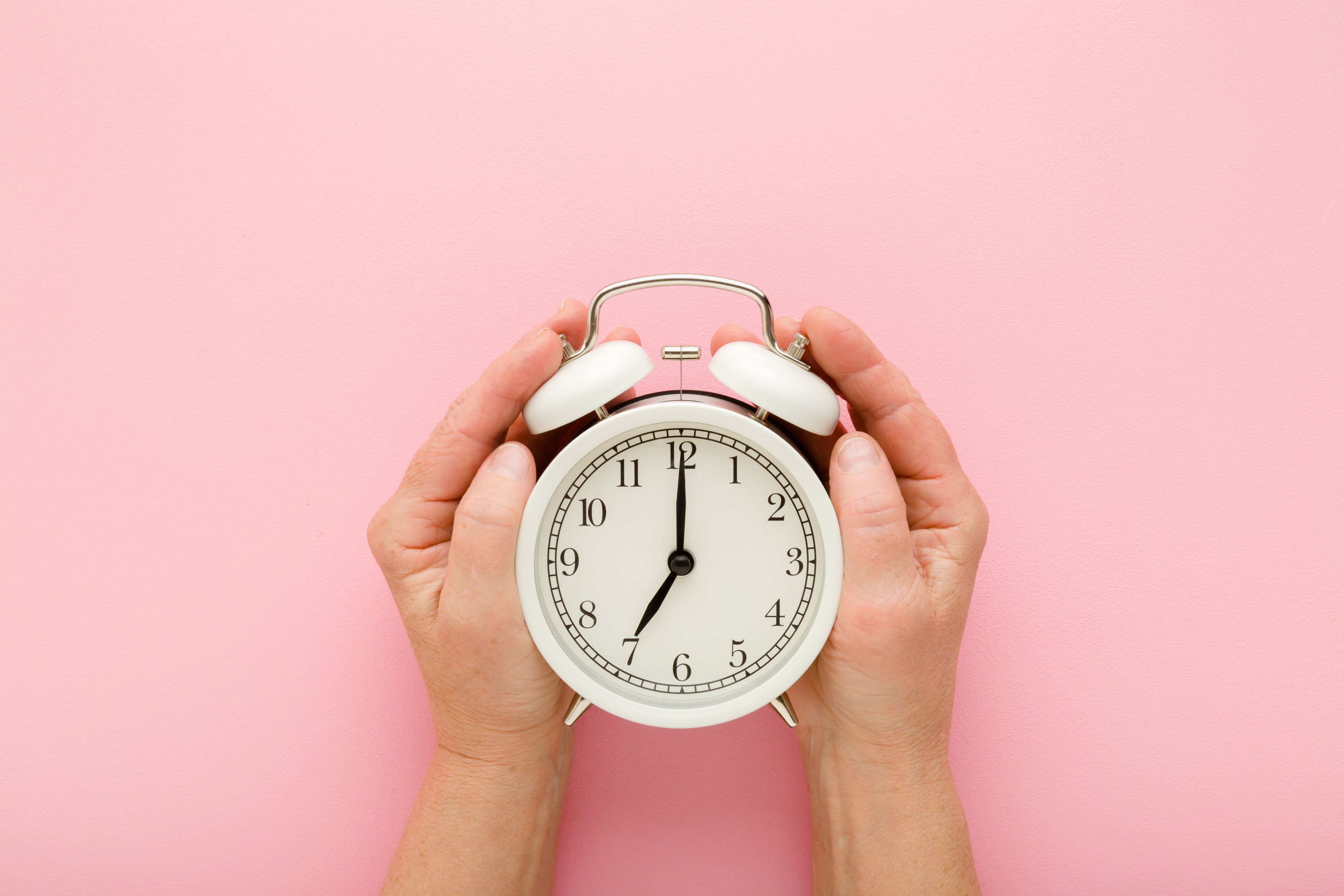 Old mature woman hands holding white alarm clock on light pink table background. Pastel color. Closeup. Point of view shot. Seven o'clock. Concept of female biological time. Top down view.