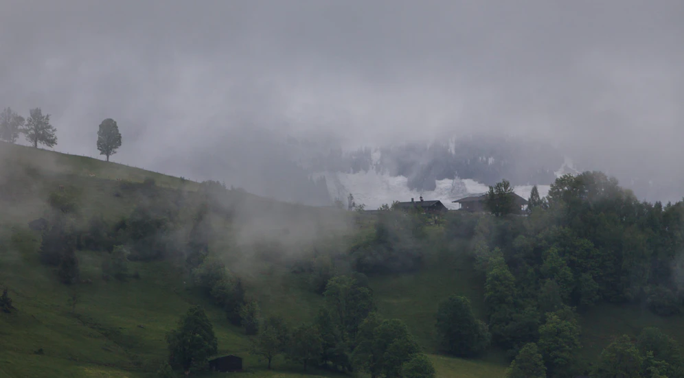 Blick auf den Maiskogel mit Bauernhäusern, dahinter ein schneebedeckter Berg, aufgenommen 2015 in Kaprun.