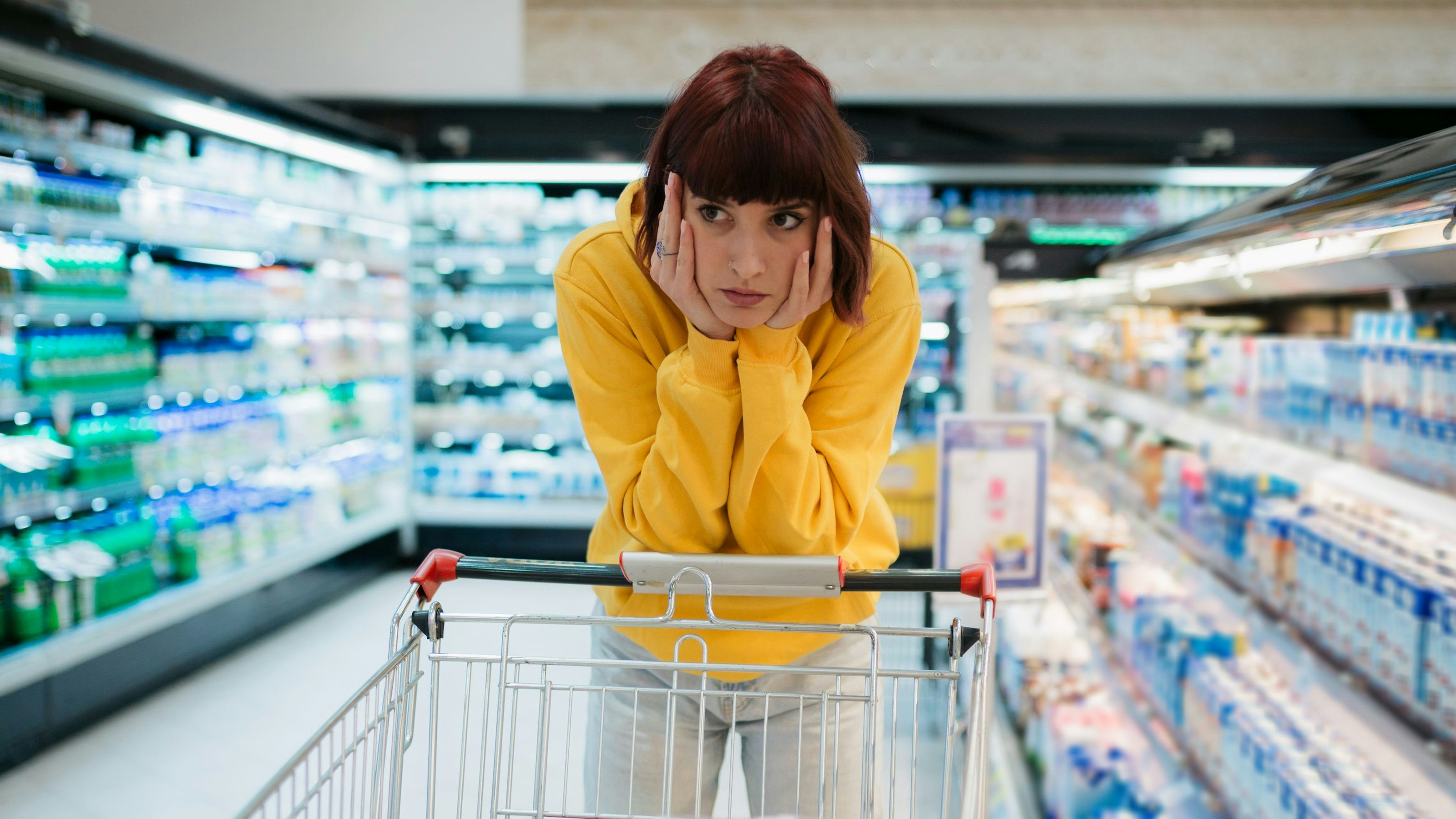 Young woman with red hair buying groceries in a local supermarket, feeling depressed by the high prices in the store and leaning on her shopping cart