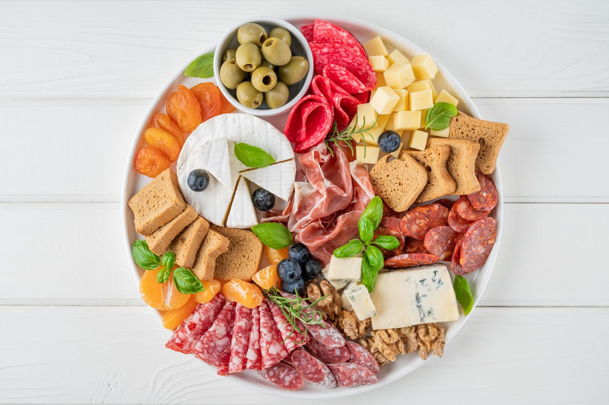 Charcuterie board with a variety of cheeses, salami, chorizzo, prosciutto, honey, grapes, nuts, olives, bread, blueberries and fresh herbs on a white wooden background. A festive snack