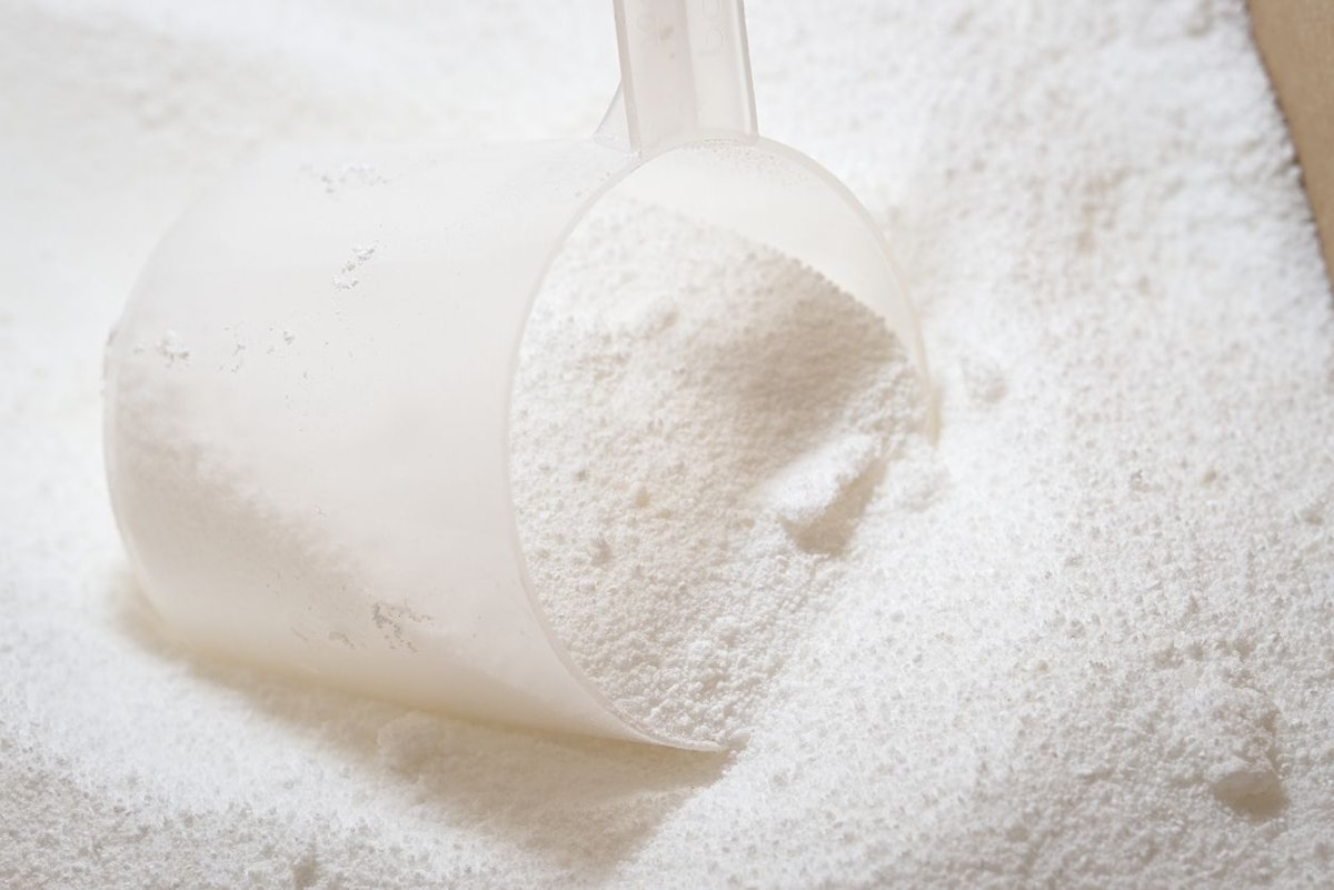 Close up of clear plastic scoop being used to measure white laundry detergent powder in a cardboard box. Bright image with macro powder detail