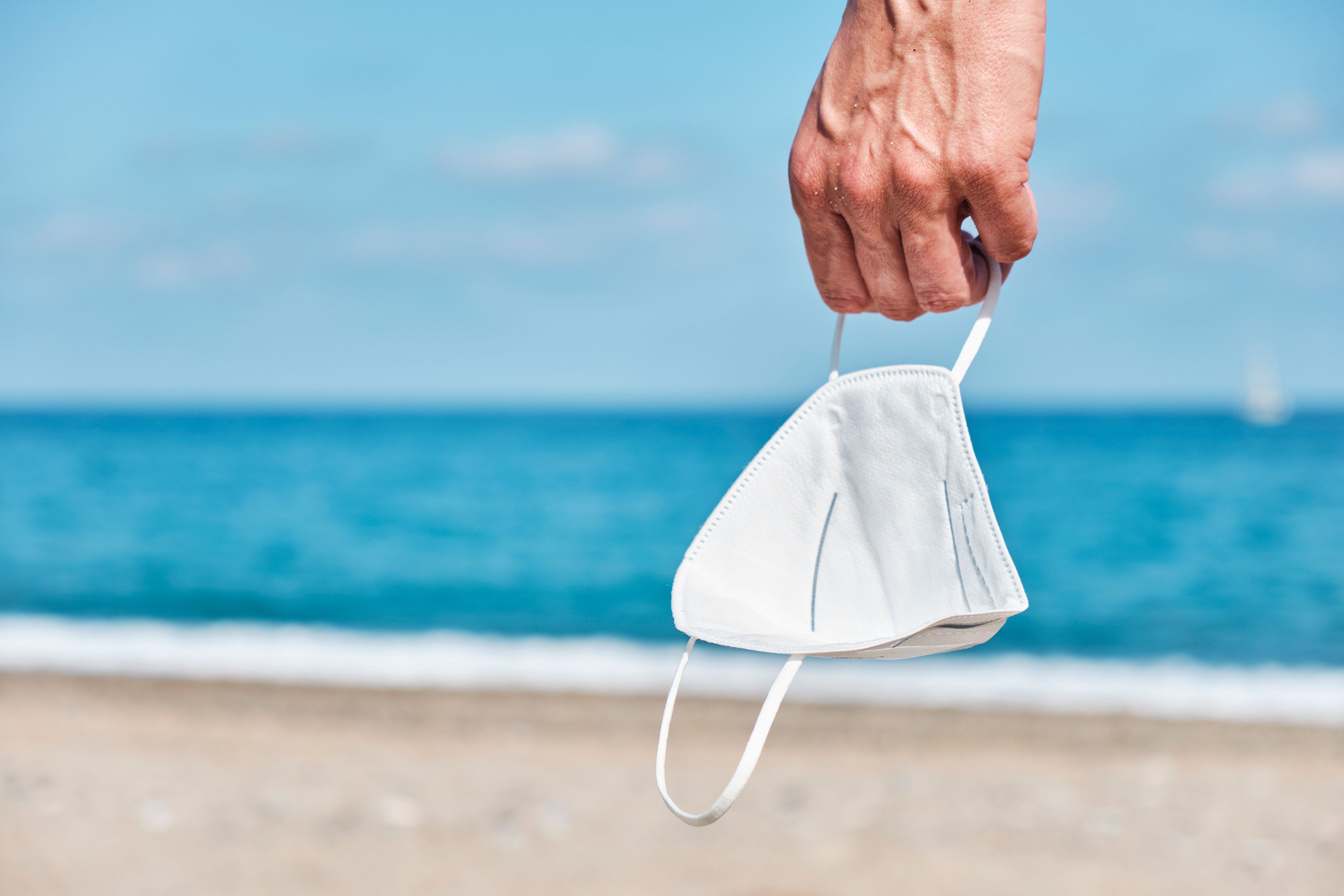 closeup of a young caucasian man, standing on the beach, holding a white face mask in his hand, as is taking a breather of wearing it, with the sea in the background