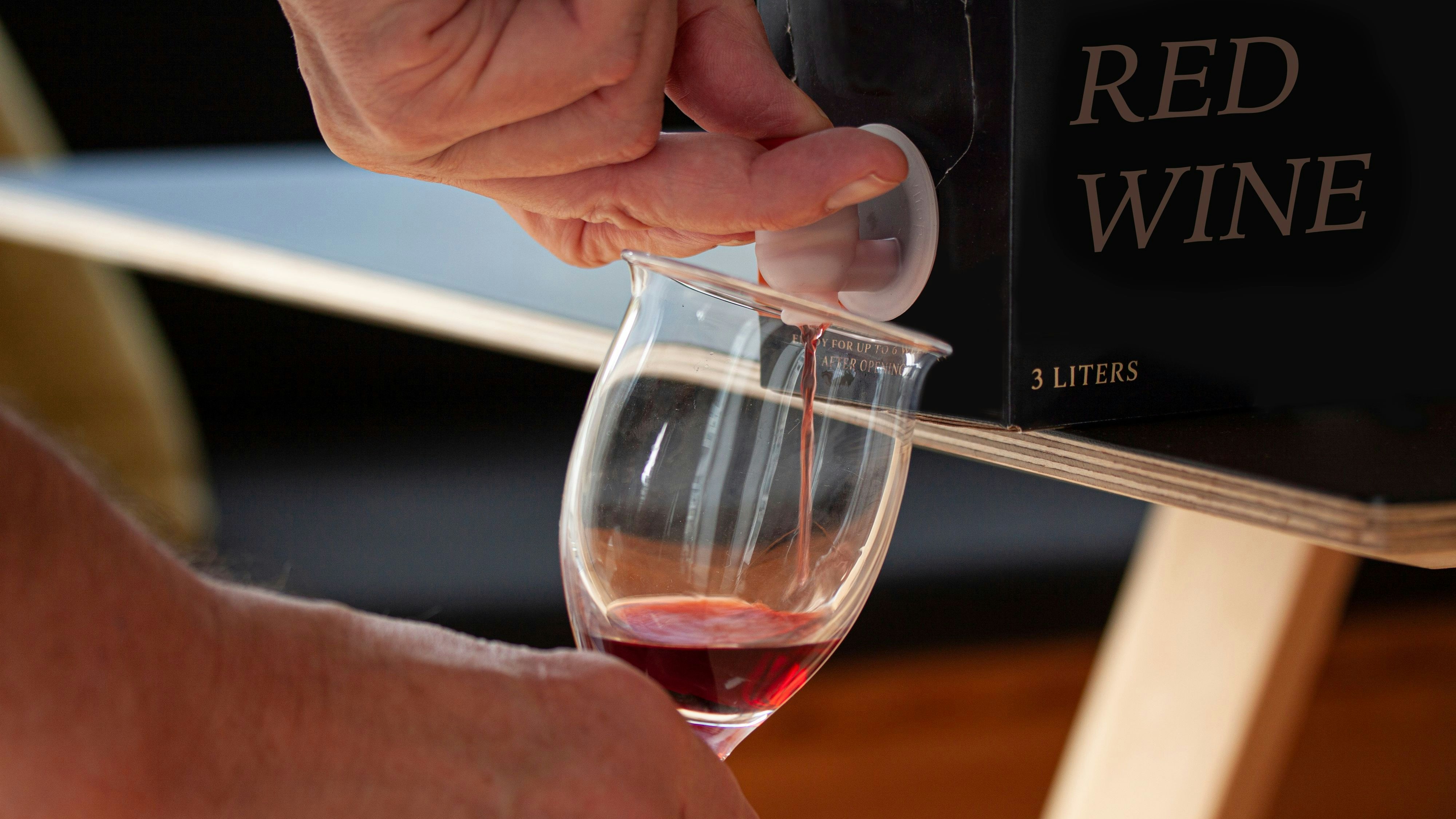 Hand pouring red wine into a glass from a BIB - cardboard bag in box with open tap standing on a table. Close up image. Fictitious brand.