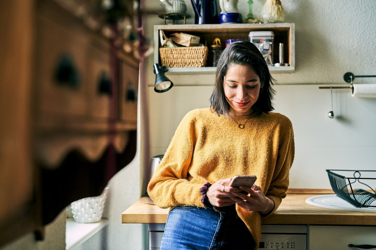 Woman leaning to kitchen counter using mobile phone. Female at home texting in cell phone.