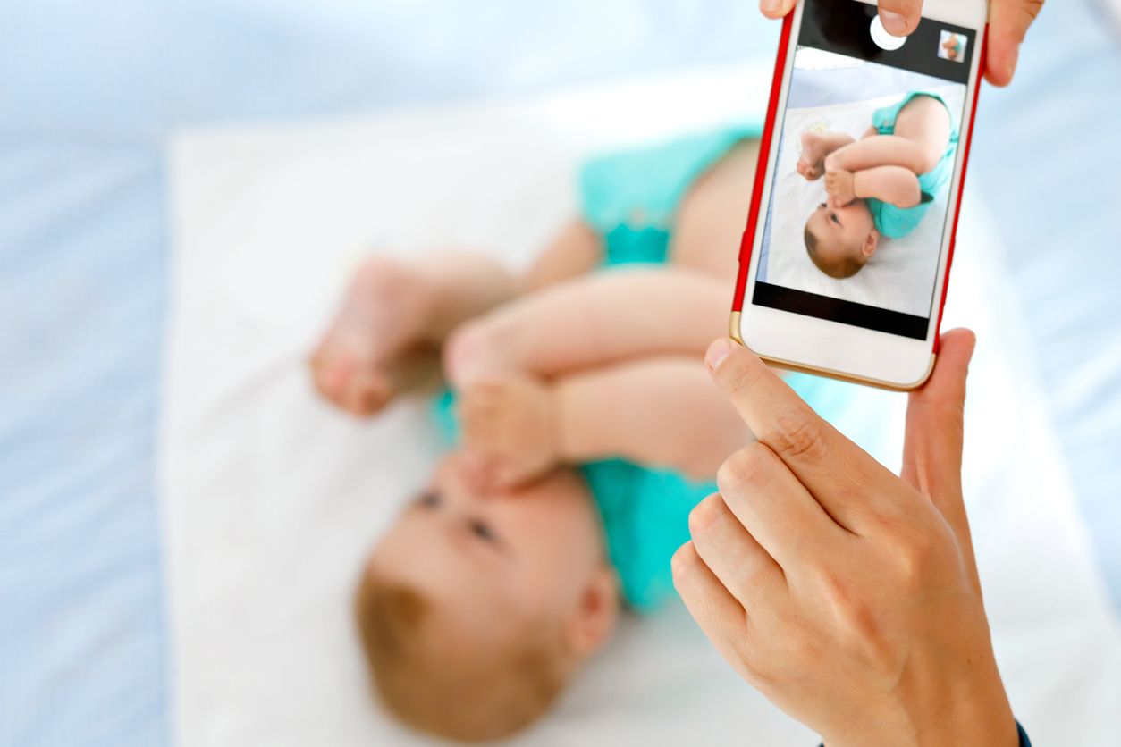 Parent taking photo of a baby with smartphone. Adorable newborn child taking foot in mouth. sucking feet. Digital family memories