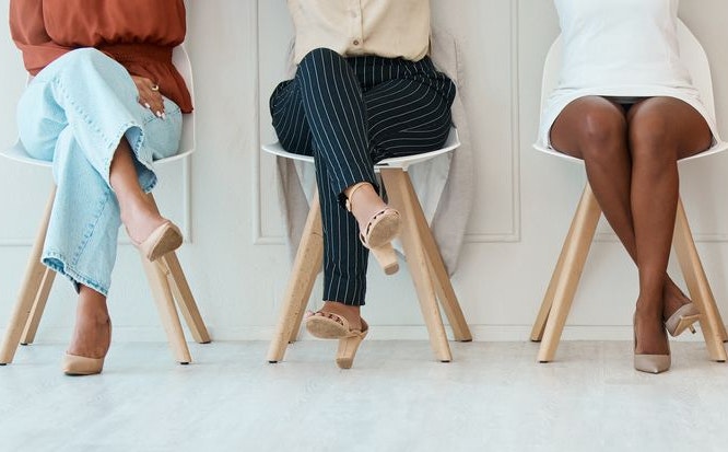 Group of five businesswomen sitting on chairs against a wall in an office at work. Legs of businesspeople sitting on chairs in a line together at work