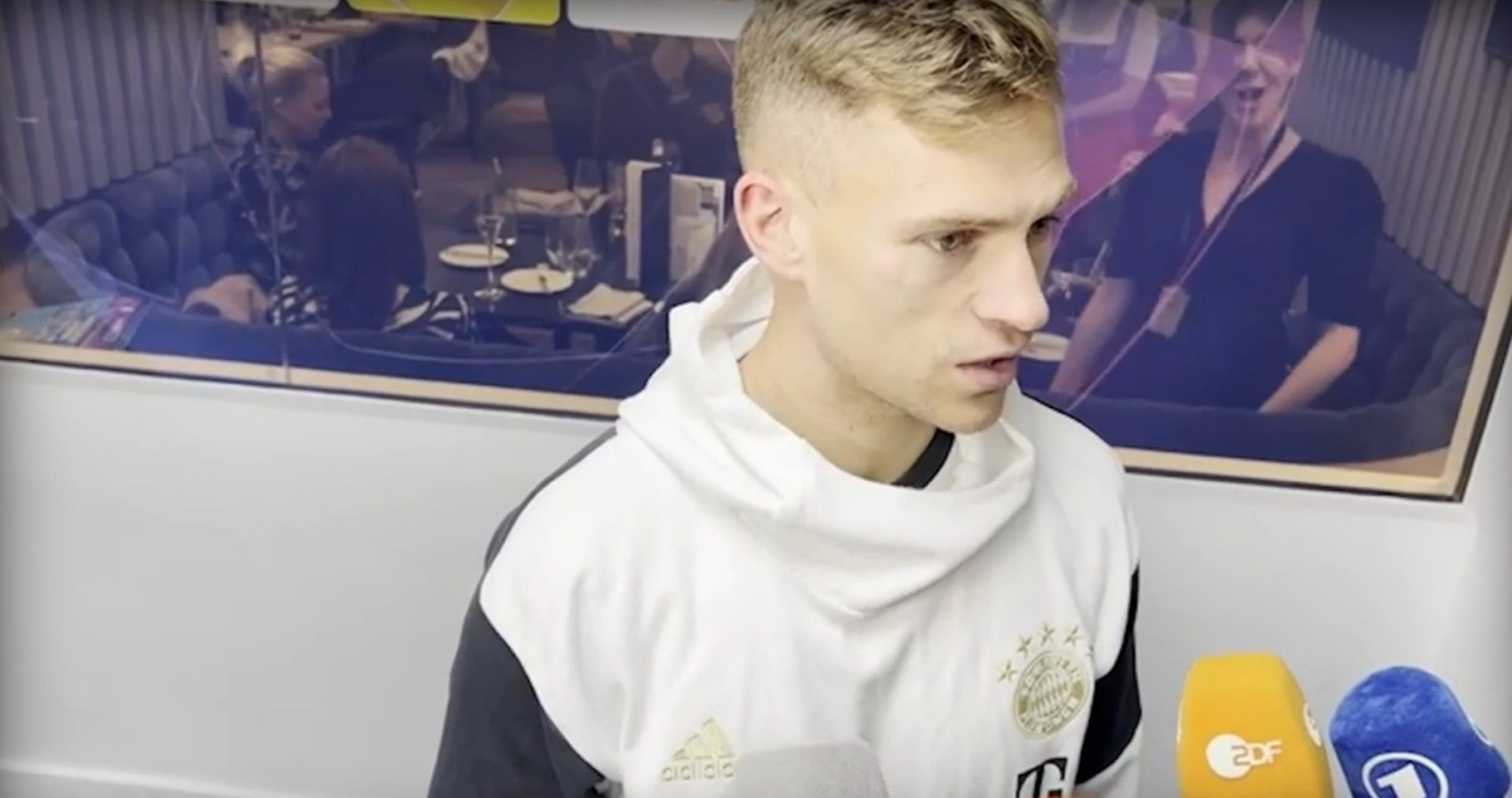 Rechts im Hintergrund wackelt die junge Frau mit ihrem Oberkörper. Im Vordergrund versucht Joshua Kimmich das Debakel zu erklären.