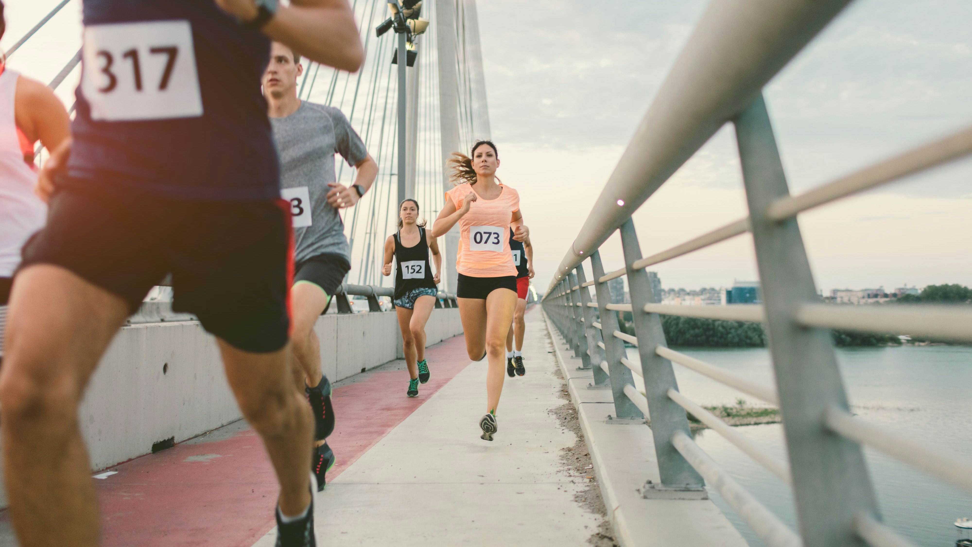 Runners running marathon in the city. They are running over the bridge at sunset. Wearing numbers on their sport clothes. Cityscape in background.