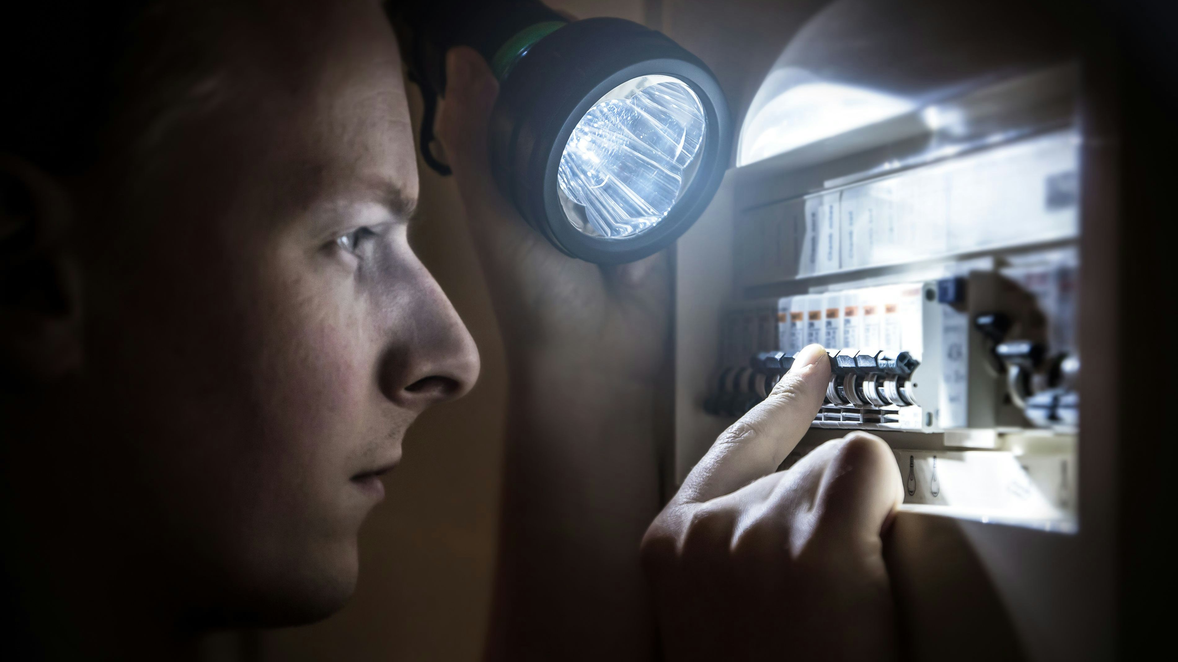 Horizontal composition photography of a young men, specialized technician or just home man, trying to restore power after electricity outage in the dark with his light. He is looking on the circuit breaker, and try to find the right button to operate. Picture shot in the dark, with a LED hand light, which gives a cold colorimetry image, and selective focus on the finger human hand and button of the circuit breaker (ON-OFF), fuse box switch. Electrical cabinet, open, with electrical circuit breaker. Side view in selective focus.