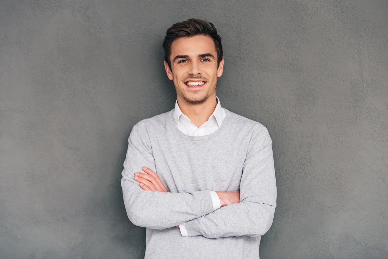 Confident young man keeping arms crossed and looking at camera with smile while standing against grey background