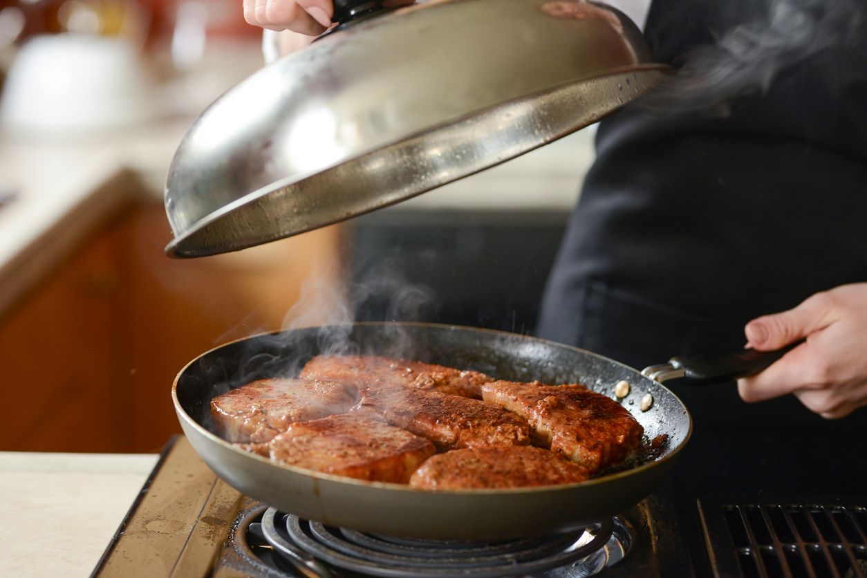 The chef in the kitchen prepares fresh pork steaks on a griddle with steam, the menu is homemade cooking culinary recipes. restaurant menu. Delicious food, recipes and cooking