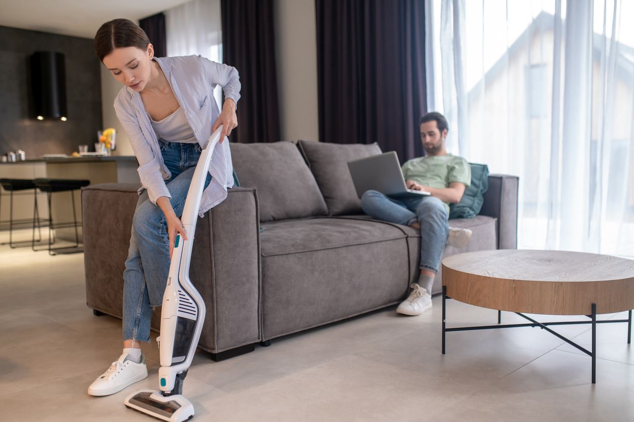 Fatigue. Tired young adult woman vacuuming crouched on sofa and man relaxed working on laptop at home during day