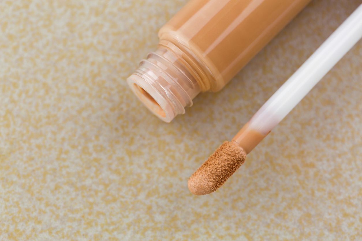 Doe-foot applicator next to tube of creamy concealer, high cover to conceal spots, blemishes, on yellow tile background