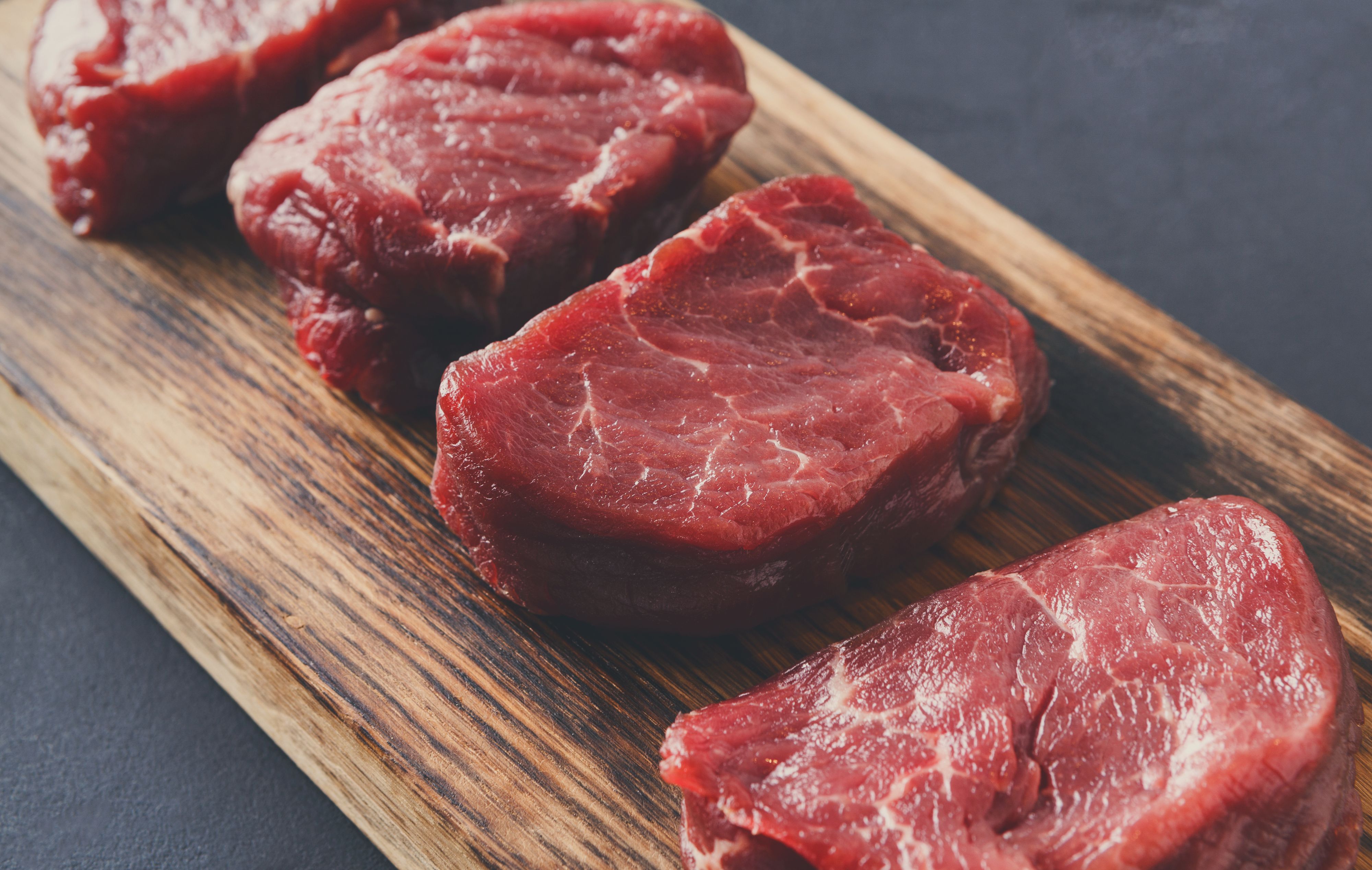 Raw filet mignon steaks. Slices of fresh beef meat arranged in a row on wooden cutting board at gray background with copy space. Organic ingredients for restaurant meals, meat texture closeup