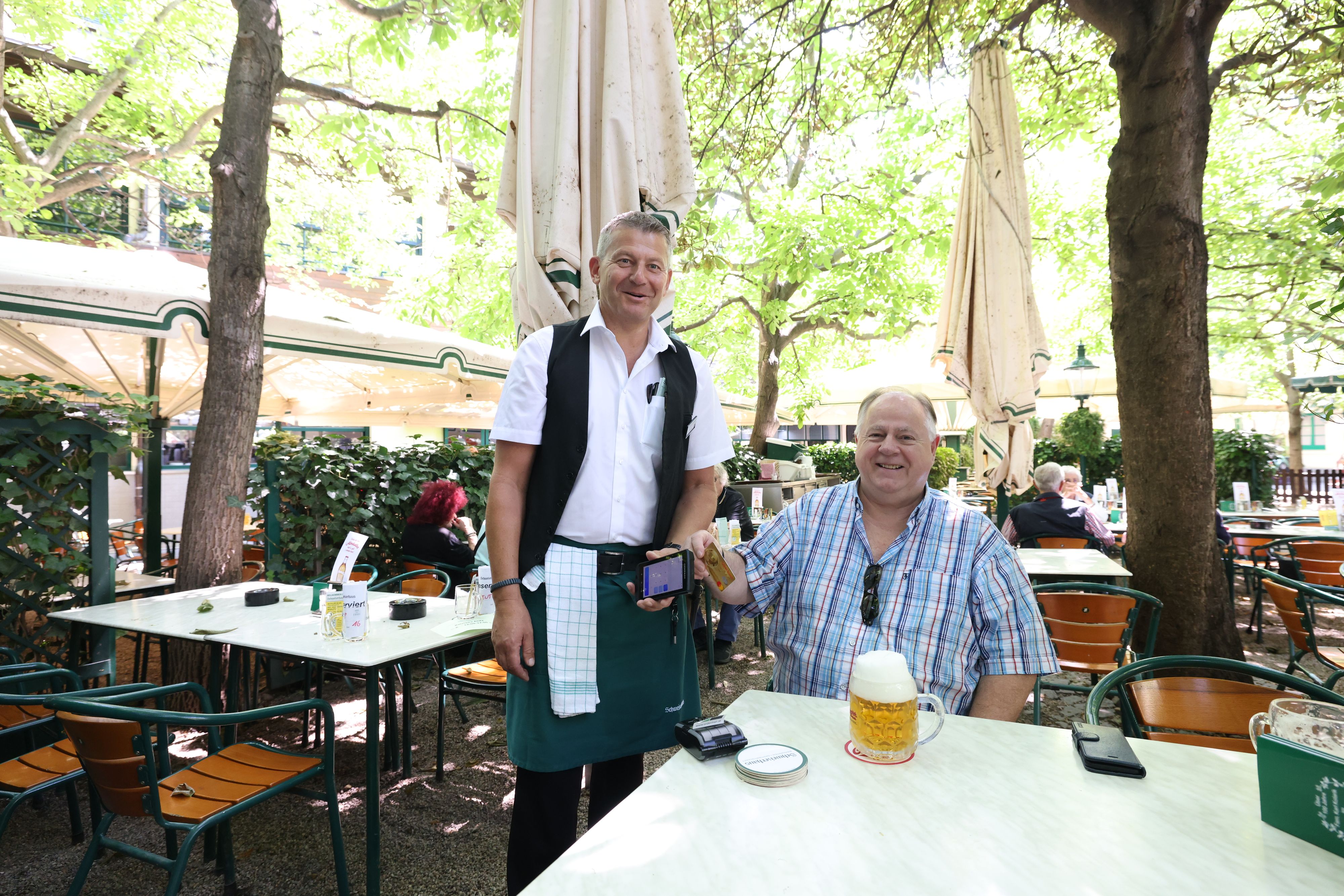 Kellner Heinz (li.) kassiert seit Saisonbeginn auch bargeldlos. Das Trinkgeld wählt Stammgast Toni direkt auf dem Bildschirm aus.