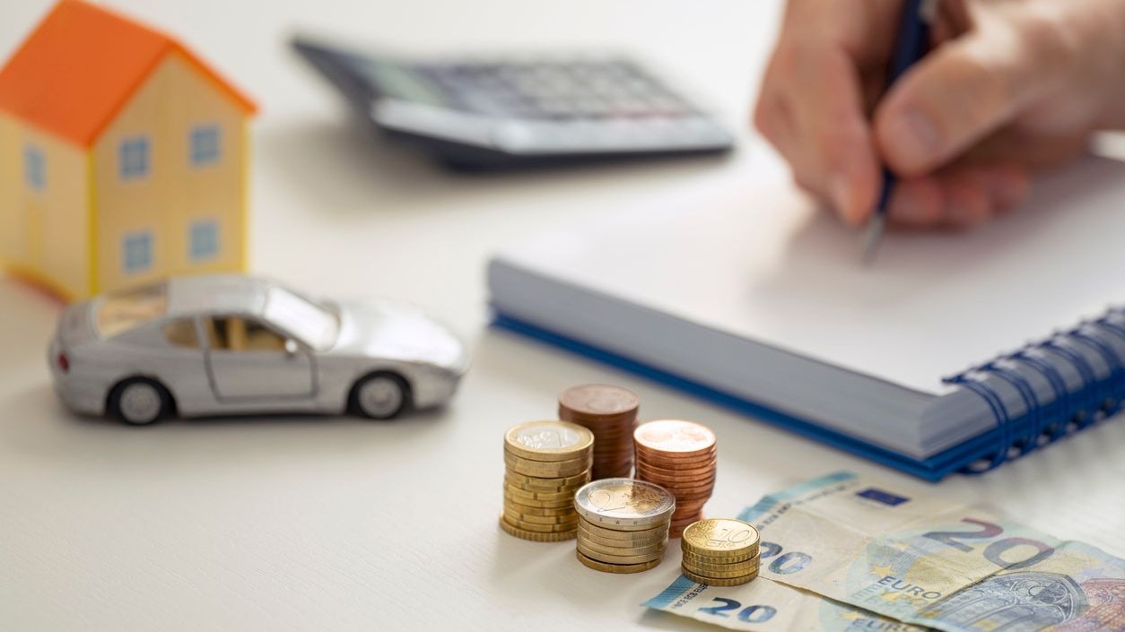 A MALE HAND WRITING IN THE NOTEBOOK NEXT TO A CALCULATOR, A LITTLE HOUSE, A TOY CAR AND SEVERAL STACKS OF EURO COINS AND NOTES. HOME ECONOMY CONTROL CONCEPT. FOCUS SELECTED.