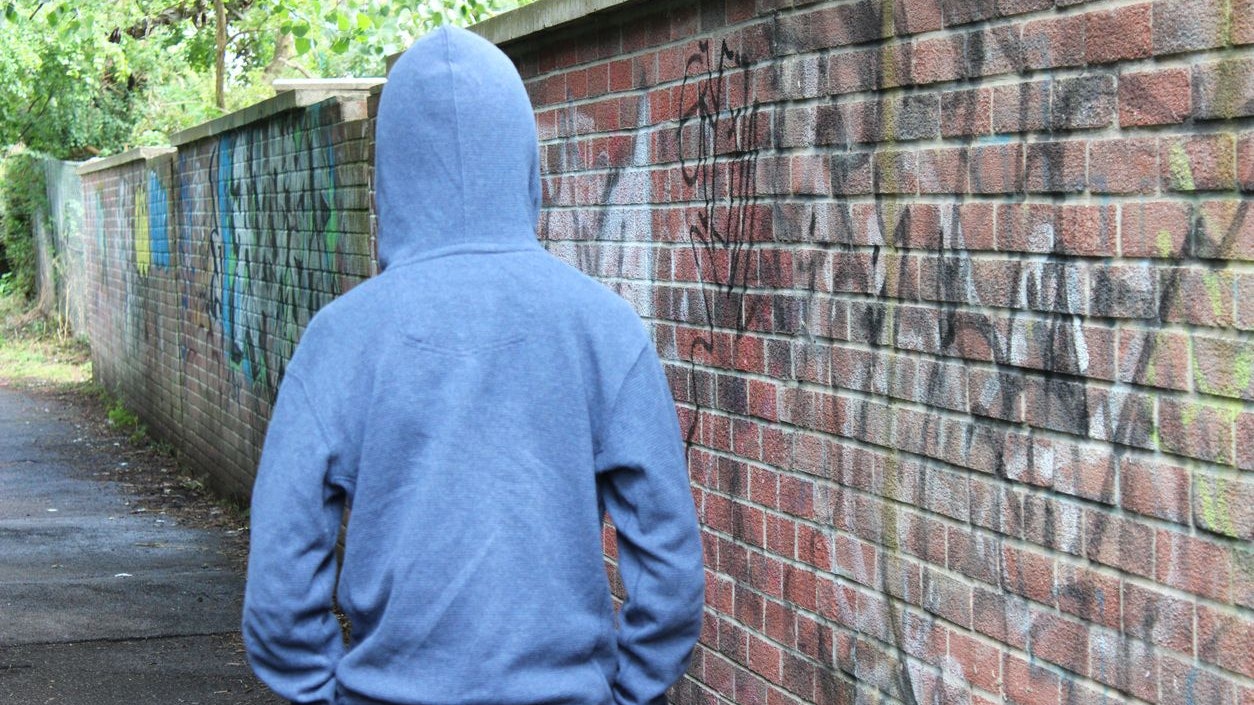 Photo showing a teenage boy / youth wearing a blue hoodie and standing beside a brick wall, in a run-down part of the city.