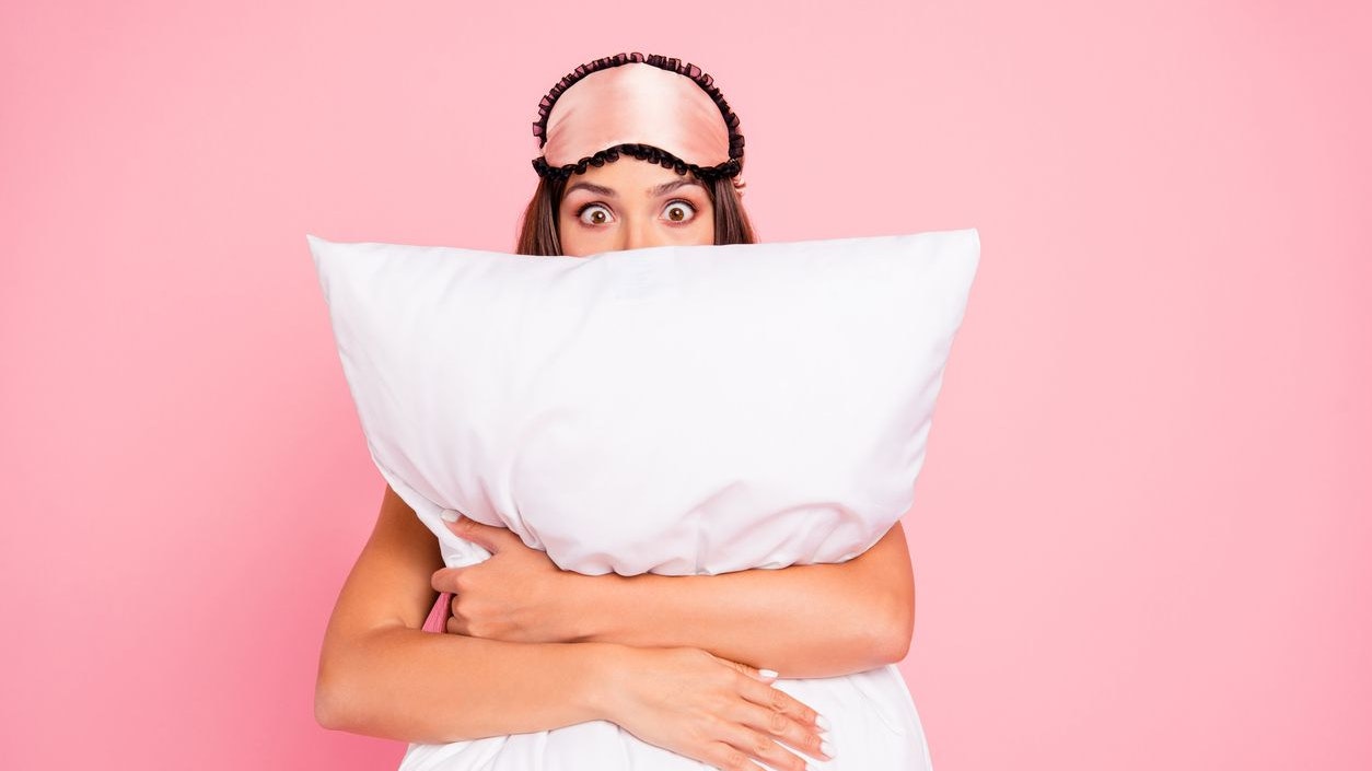 Young gorgeous shocked lady wearing eye mask, hugging pillow, hiding. Isolated over pink pastel background