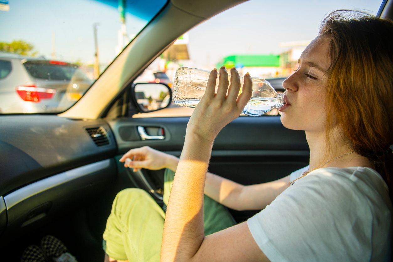 The attractive long-haired Caucasian young girl drinking water and resting at the passenger seat in the car during the roadtrip in the USA. Summer vacations, the sunny hot day on the city street.