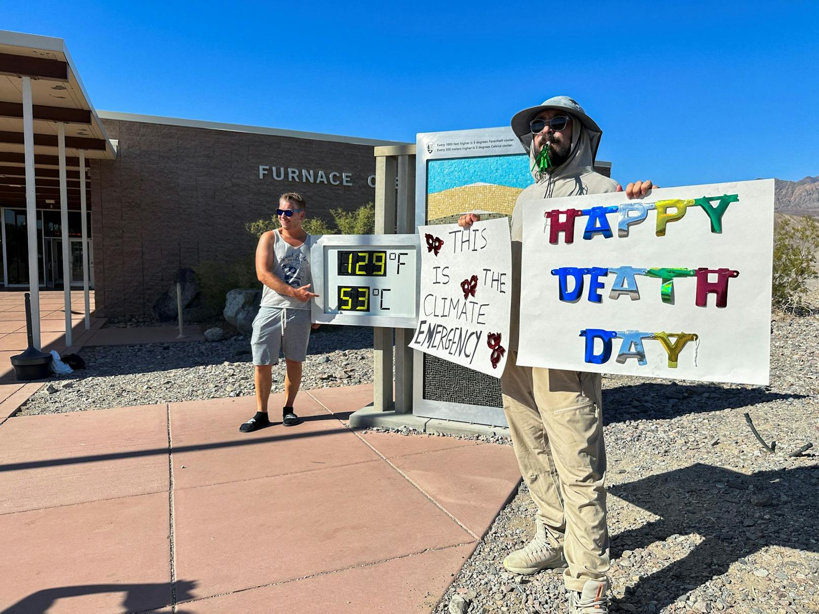 Death Valley: Während im Vordergrund ein Mann auf die Klimakrise aufmerksam will ("Das ist ein Klima-Notfall. Einen fröhlichen Todestag"), posiert ein Mann im Hintergrund vor der Temperaturanzeige.