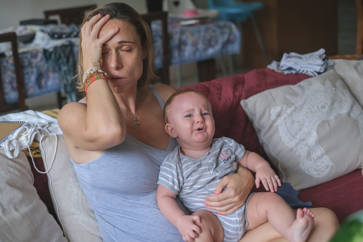 Front view shot of a stressful Caucasian mother holding her crying baby boy with one hand. They're both sitting on the sofa in the living room.