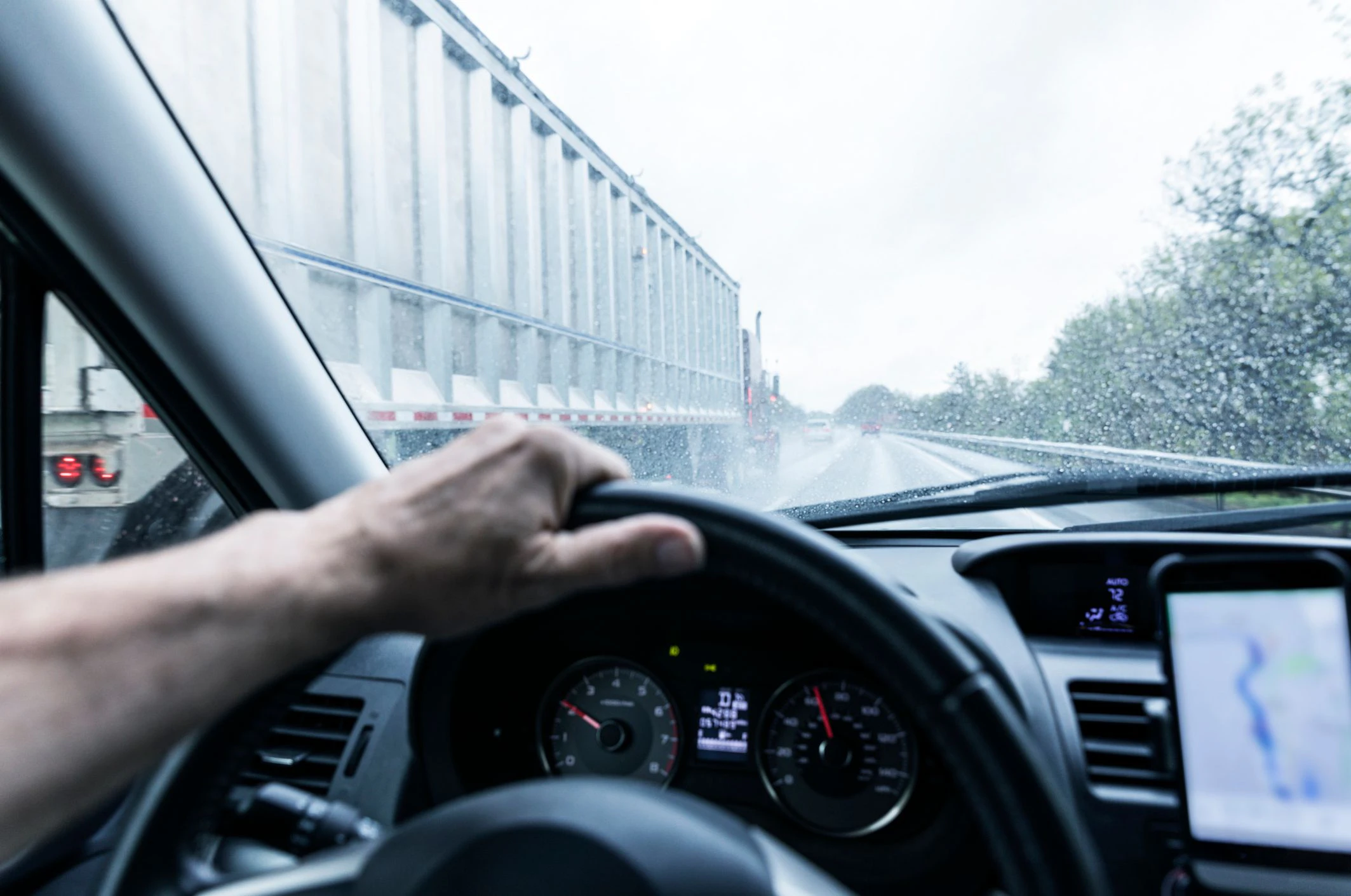 Car driver point of view steering and looking through windshield on a stormy rainy day while a semi tractor trailer truck speeds past in the left lane on a rural road stretch of expressway highway. The windshield wipers are trying to keep up with the water road spray kicked up by the truck's tires. A generic smart phone display mounted on the dashboard running a mobile navigation app is providing directions. -- (Note to Inspection Team - This is a replacement for an earlier "revisable" image in this batch.)
