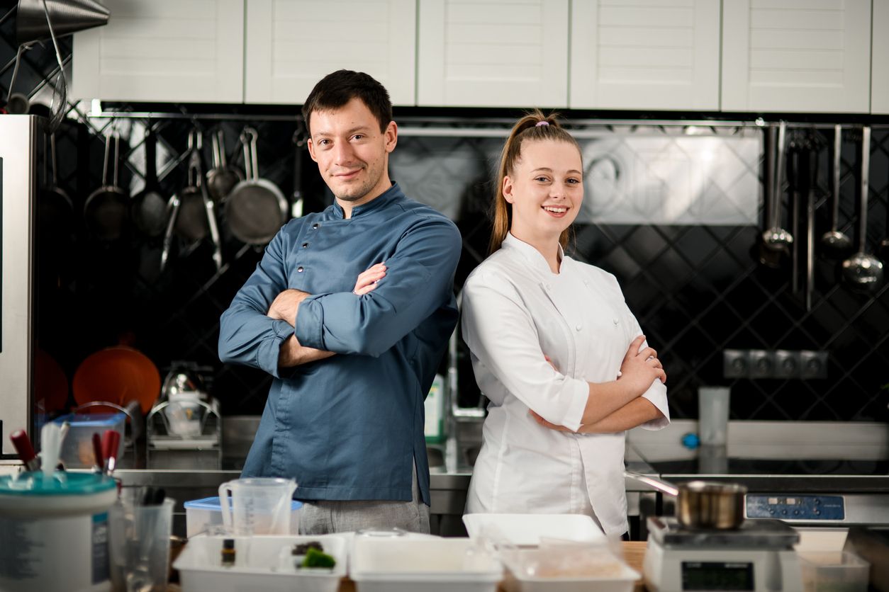 young beautiful smiling man and woman chefs stand in the kitchen with crossed arms. Blurred background