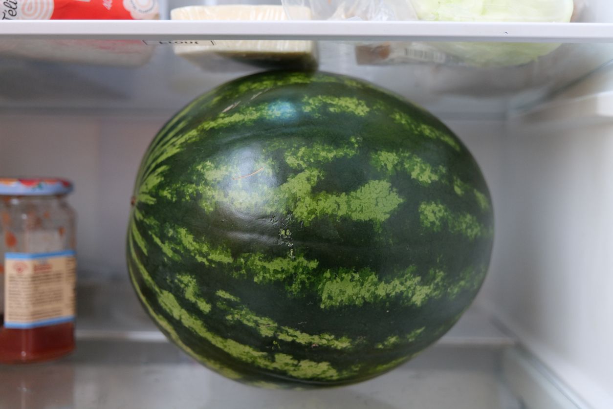 A green and whole watermelon is on the shelf of a refrigerator. There is a bottle of jam next to it and some other food above it.