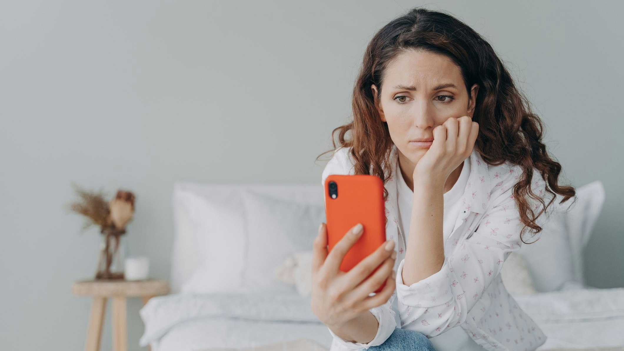 Stressed caucasian girl is reading morning news looking at smartphone display. Young woman is chatting on phone or browsing internet at home in her bedroom. Lady is doom scrolling.