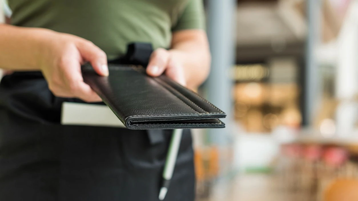 Cropped image of woman waitress in apron hold out bill folder to cafe visitor of cafe or restaurant.