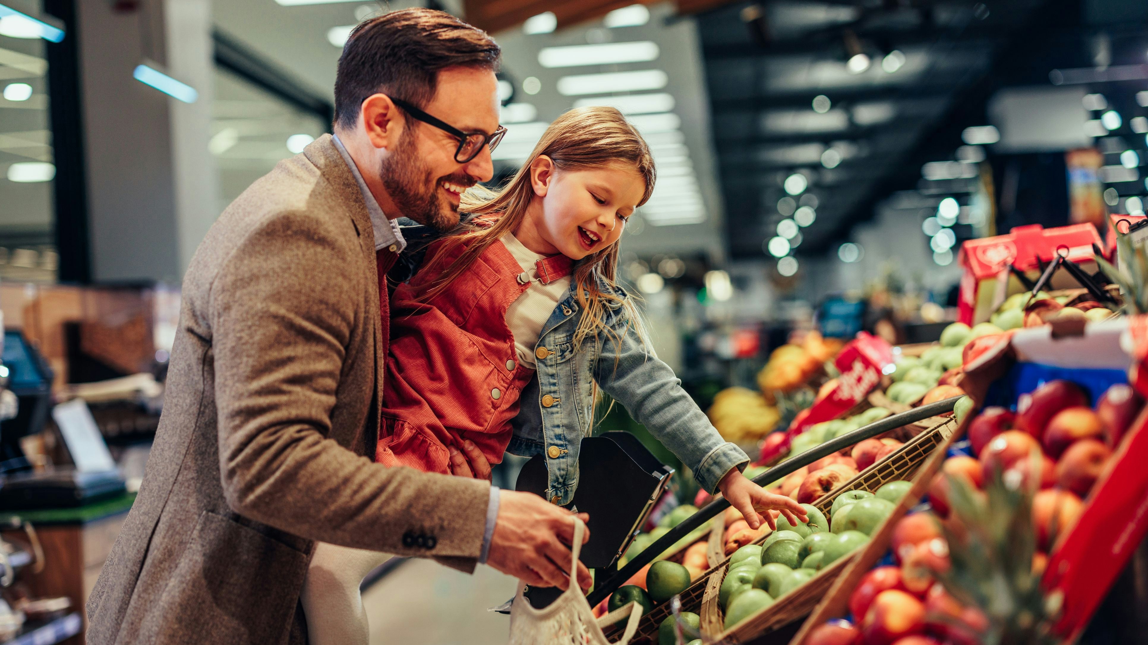 Little girl is buying groceries in the supermarket with her father. He carrying her and picking together fresh fruits