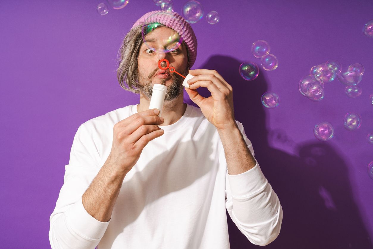 Carefree middle aged man blowing soap bubbles against purple background.