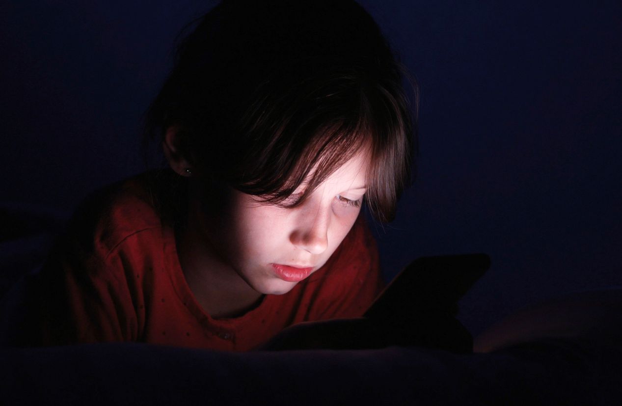 Caucasian girl lying in bed playing tablet in social internet in dark blue light under blacket. Childhood, communication and technology concept