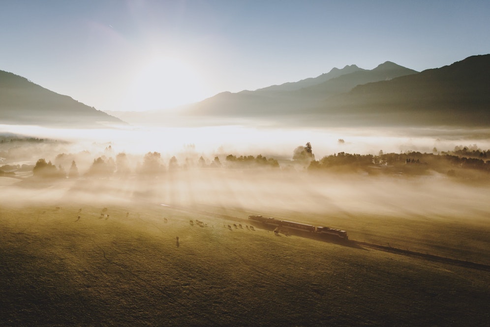 Sonnenstrahlen erhellen die mit Nebel bedeckte Landschaft mit einer Herde Milchkühe und einer Zuggarnitur der Pinzgauer Lokalbahn. Aufgenommen am 29. September 2023 in Kaprun.