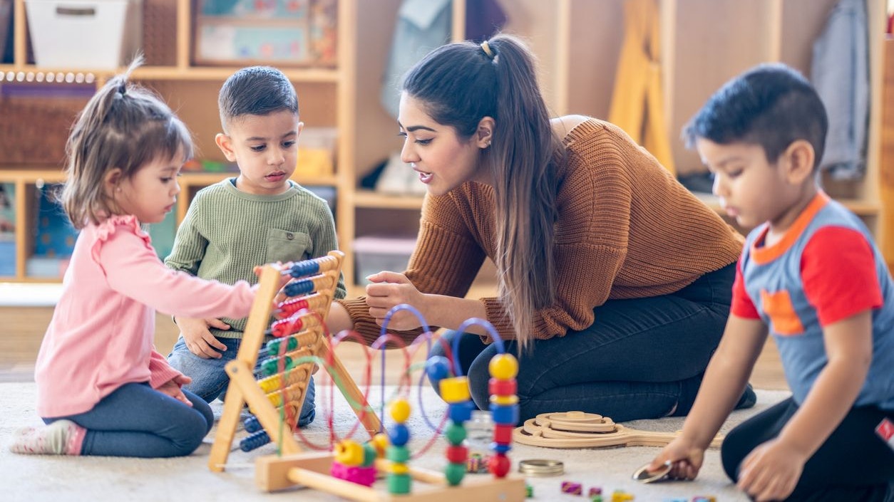 A female Kindergarten teacher of Middle Eastern decent, sits on the floor with students as they play with various toys and engage in different activities.  They are each dressed casually as they learn through their play.