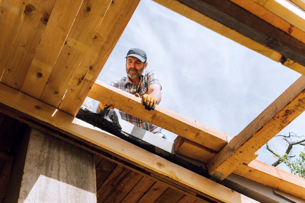 Male carpenter crouching while using hammer on wooden roof beam of incomplete residential structure at site