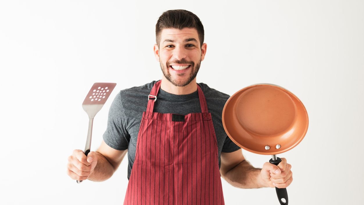 Hispanic guy in his 20s with an apron is happy and ready to start cooking. Young guy holding a frying pan and a spatula