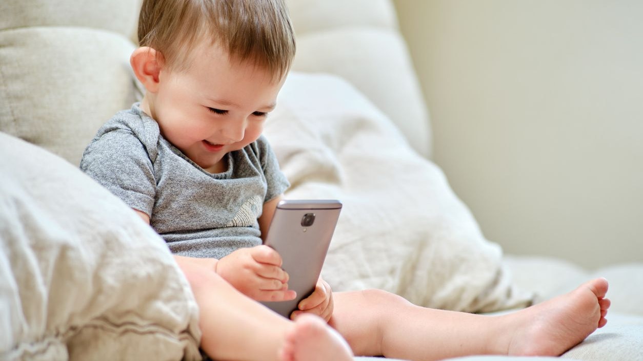 Happy toddler baby boy is sitting with a phone on the sofa in the living room. Child with a smartphone in his hands on the bed